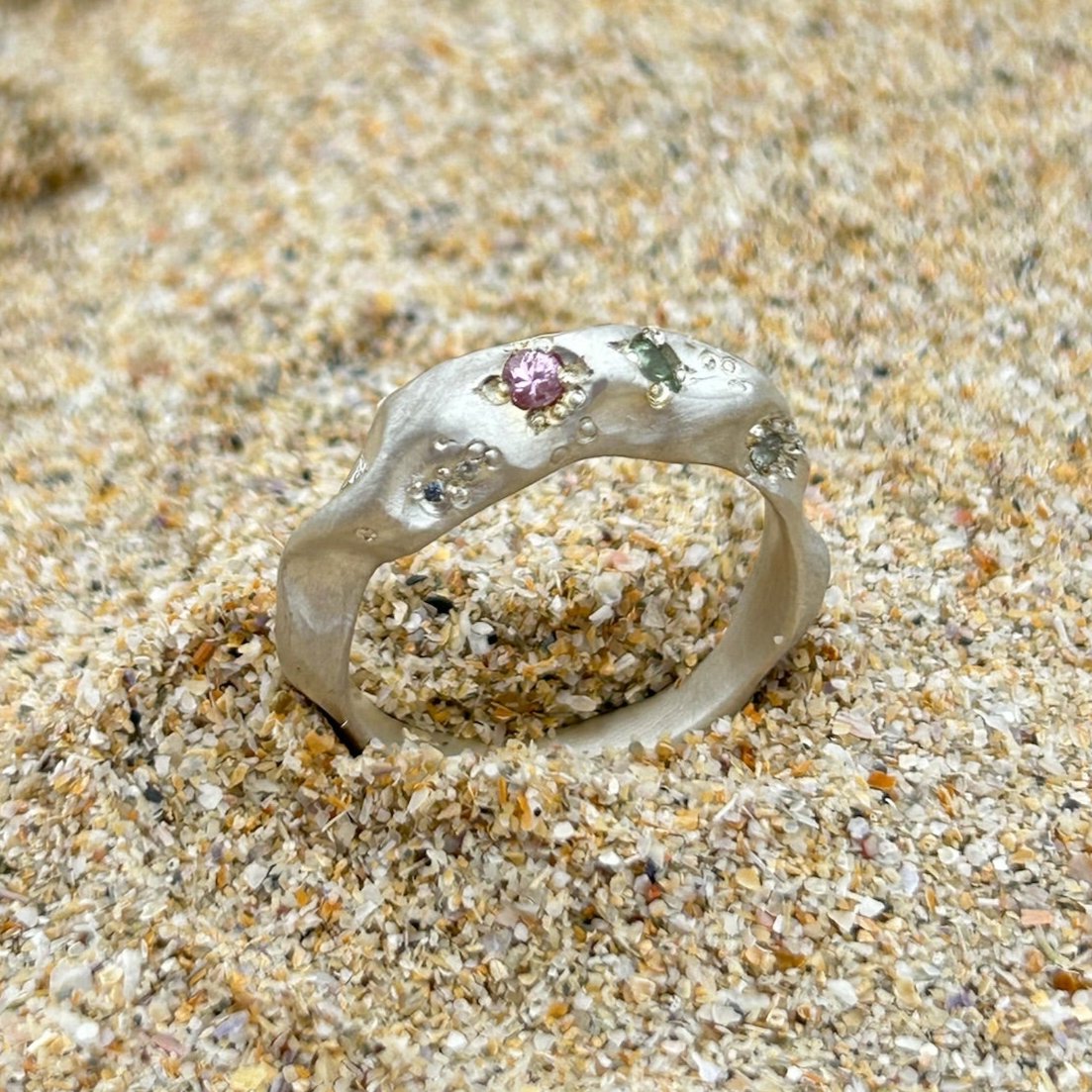 A silver ring set with sapphires in Pink, blue and green sits on a sandy beach