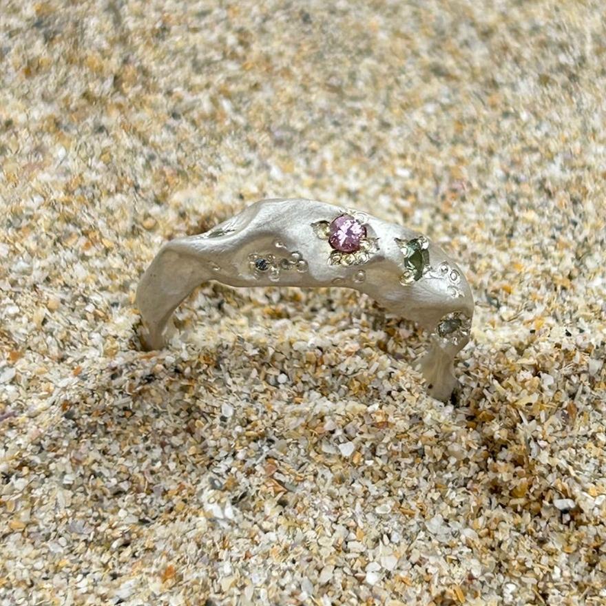 A sterling silver ring set with coloured sapphires sits on a sandy beach