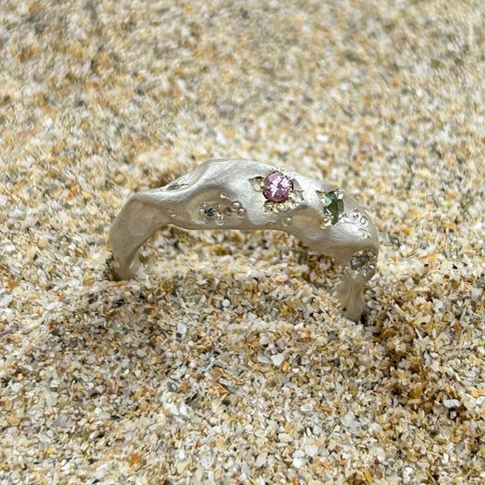 A sterling silver ring set with coloured sapphires sits on a sandy beach