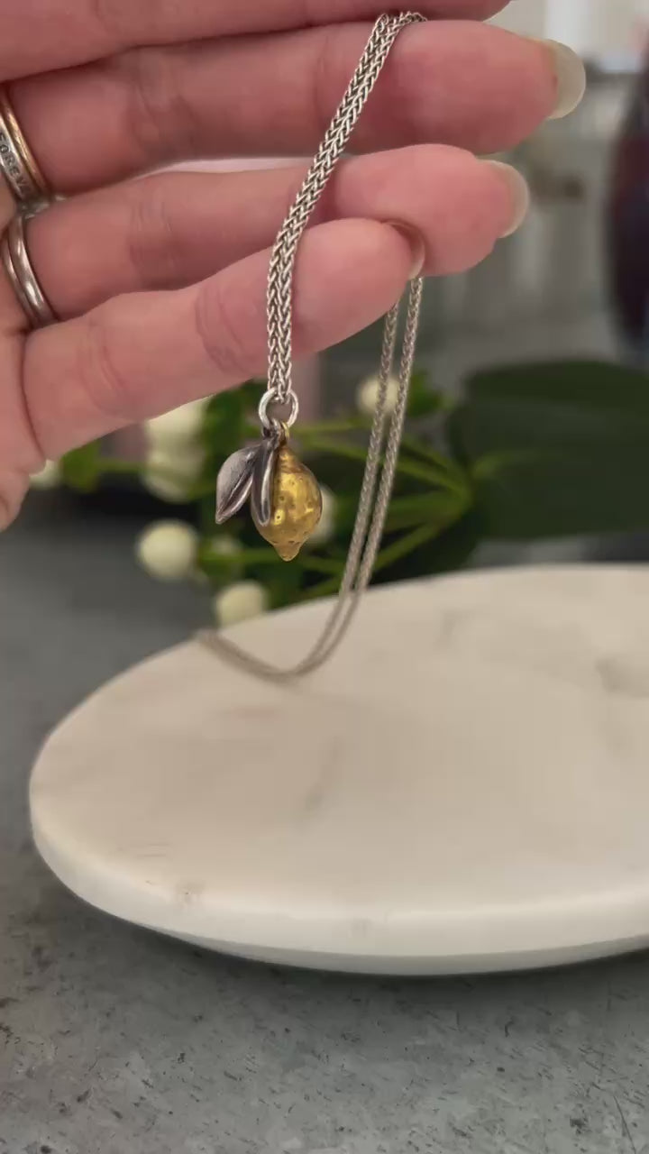 Video shows a hand holding a handcrafted Lemon pendant, the lemon is plated in 18ct gold with oxidised silver leaves, hanging from a silver chain.