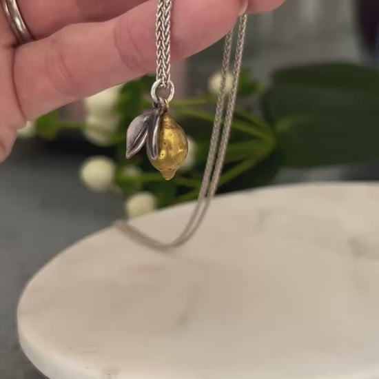 Video shows a hand holding a handcrafted Lemon pendant, the lemon is plated in 18ct gold with oxidised silver leaves, hanging from a silver chain.